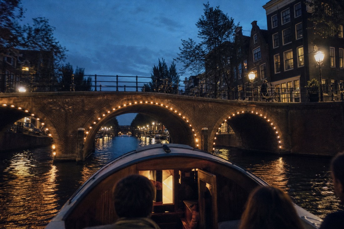 An evening canal cruise boat passing under a lit Amsterdam bridge, no text, no logos, no people