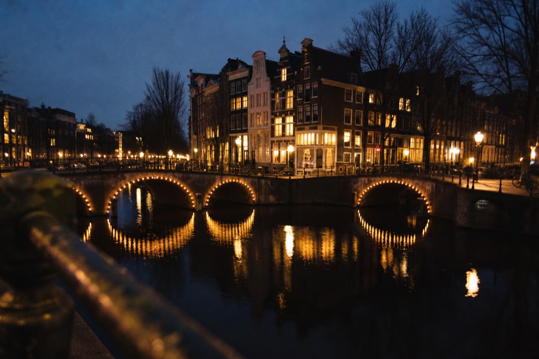 Amsterdam canal ring at dusk with lit bridges reflecting on the water, no text, no logos, no people.
