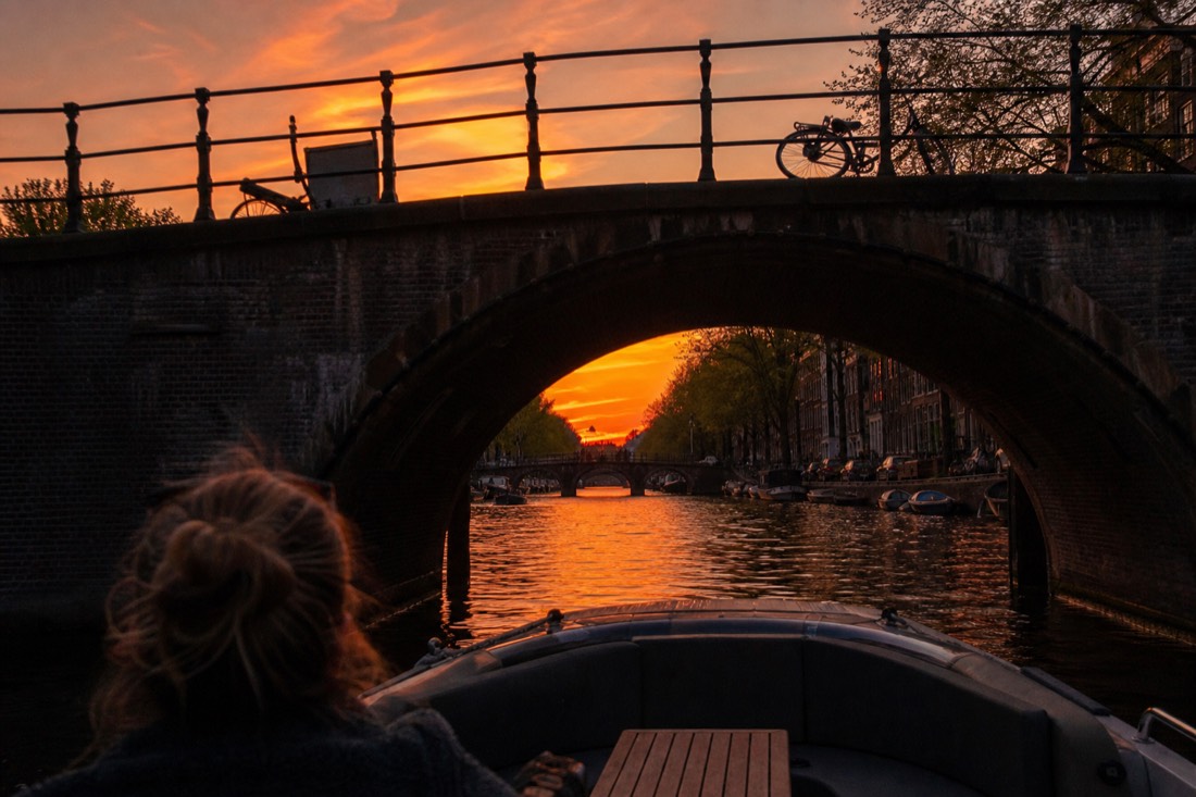 An open electric boat passing under a classic Amsterdam canal bridge at sunset, no text, no logos, no people.