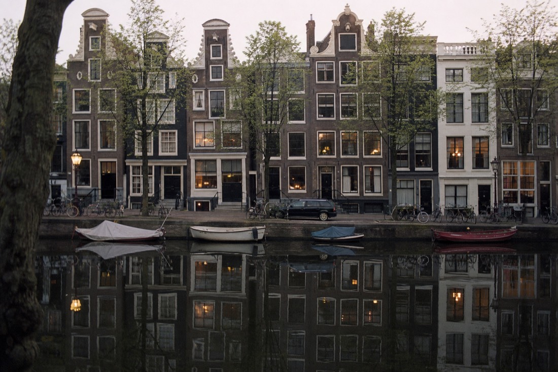 Row of narrow Amsterdam canal houses reflected in still evening water, no text, no logos, no people.