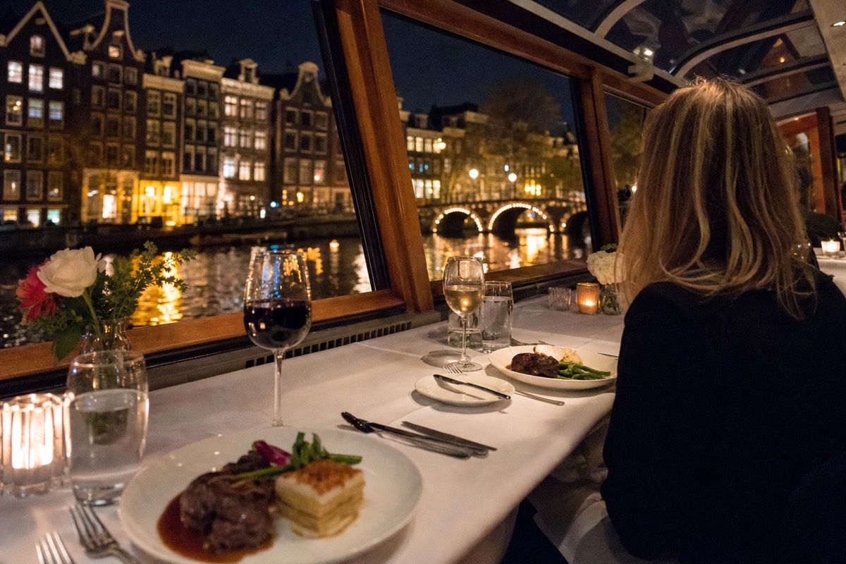 A formally set three-course dinner table aboard an enclosed Amsterdam canal cruise boat at night, no text, no logos, no people